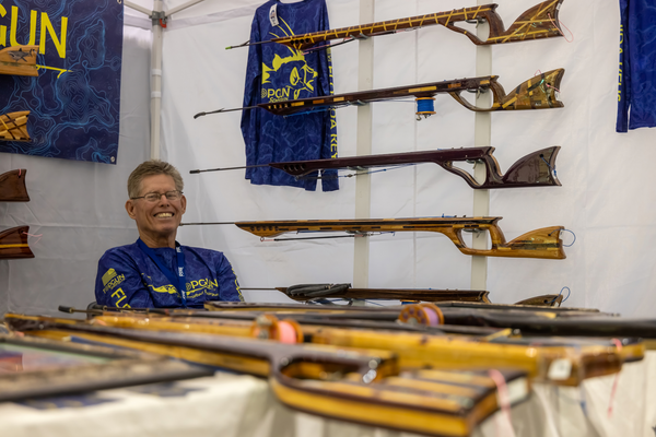 A Topgun Speargun craftsman smiles beside his handcrafted wooden spearguns at the Florida Saltwater Fishing Expo.