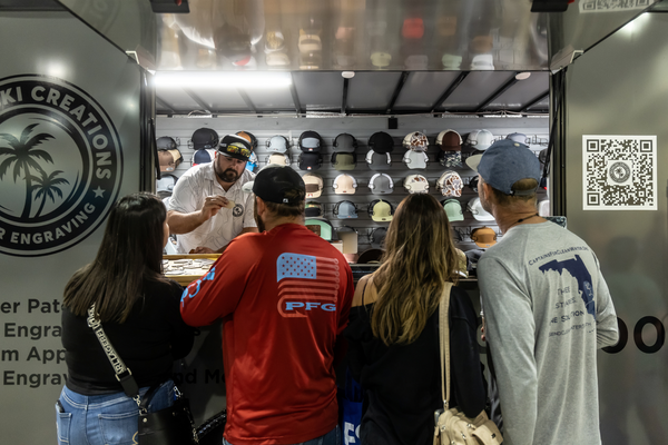 A hat customization trailer offers laser engraving on ball caps while a crowd browses the wall of options at the Florida Saltwater Fishing Expo.