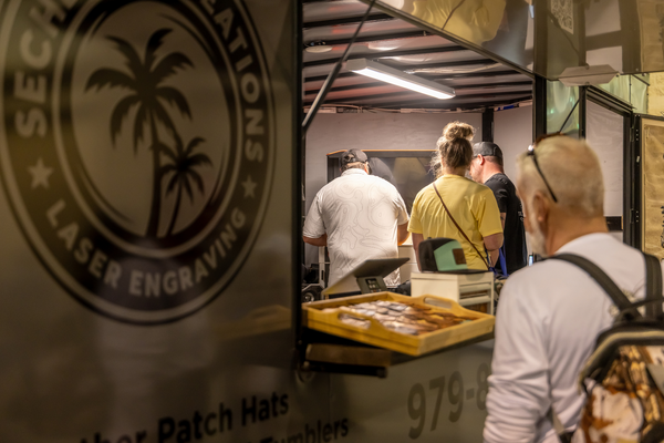 Customers wait at a laser engraving trailer offering custom leather patch hats and tumblers at the Florida Saltwater Fishing Expo.