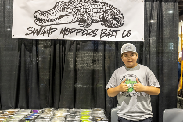 A grinning Swamp Moppers Bait Co. team member throws up a double thumbs-up in front of the shop's gator-logo banner at the Expo.