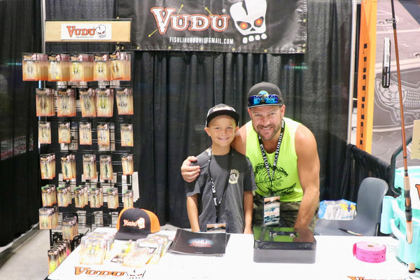 A proud dad and his son share a big smile at the Vudu lures booth during the Florida Fishing Expo, surrounded by rows of soft-plastic baits.