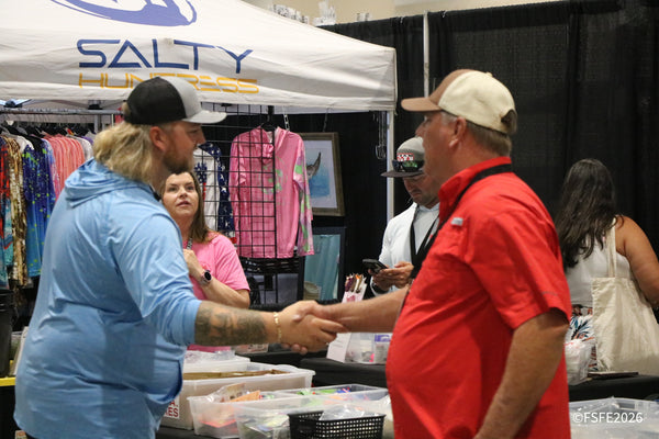 Two attendees share a warm handshake at the Salty Huntress apparel booth during the Florida Fishing Expo.