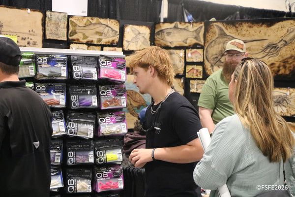 Shoppers browse Q8 Superbait soft plastics beneath a wall of hand-burned wood fish art at the Florida Fishing Expo.