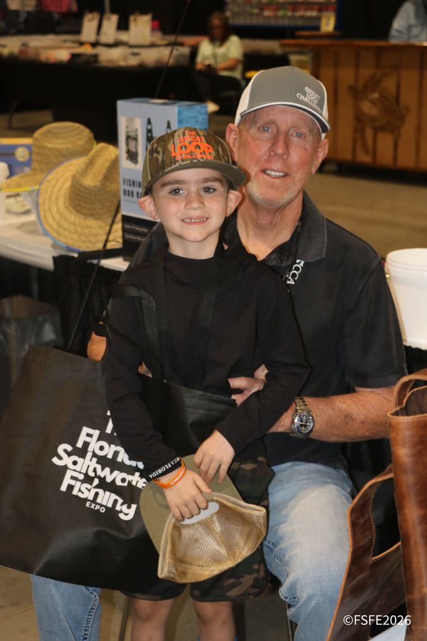 A smiling grandfather and young boy pose together holding a Florida Fishing Expo tote bag and hat.