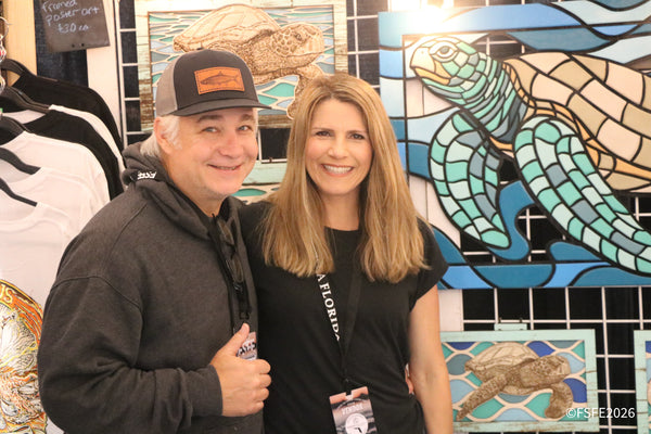 A smiling vendor couple poses in front of colorful sea turtle mosaic art at the Florida Fishing Expo.