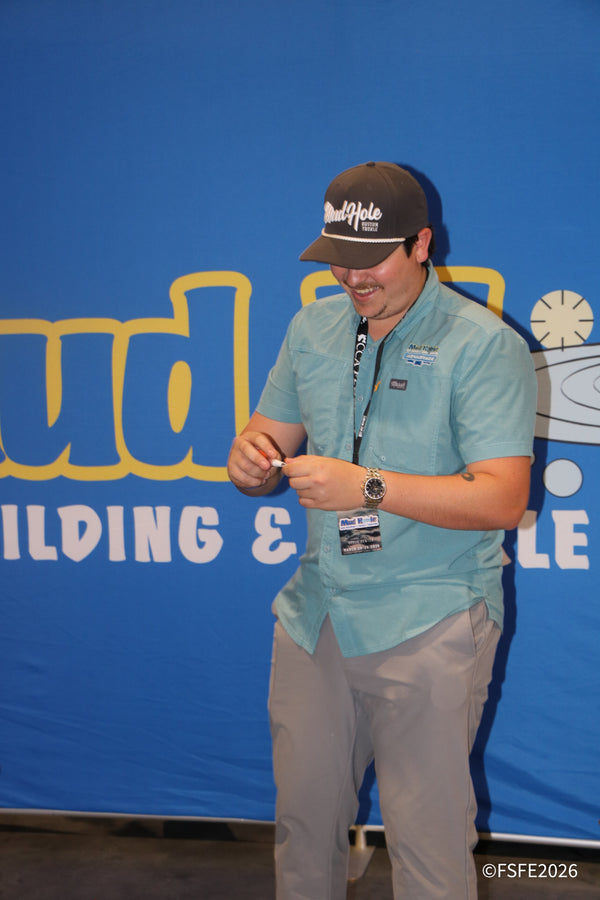 A smiling Mud Hole Custom Tackle team member signs autographs in front of the brand's rod-building banner at the Florida Fishing Expo.