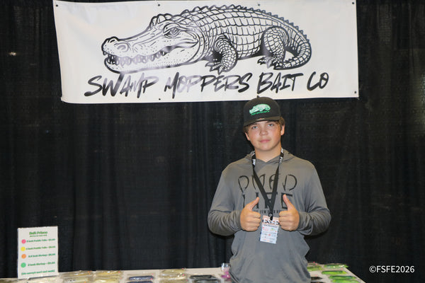 A young Swamp Moppers Bait Co. team member gives two thumbs up beneath the booth's bold alligator logo at the Florida Fishing Expo.