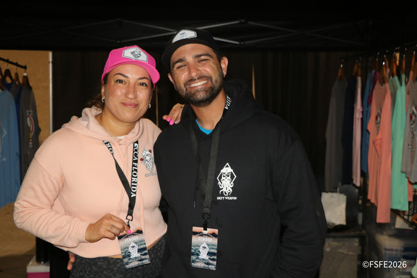 A vendor couple flashes big smiles at the Salty Weapon apparel booth, racks of fishing tees lining the wall behind them at the Florida Fishing Expo.