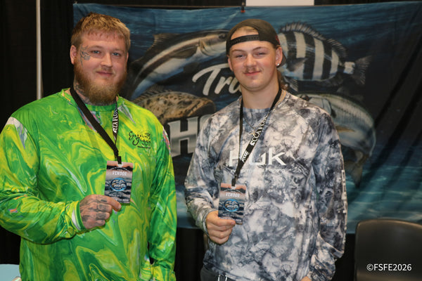 Vendors from Truly Hooked Custom Jigs, sporting Huk gear, smile and hold up their badges at the Florida Fishing Expo.