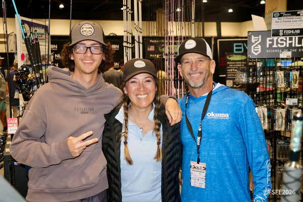 The smiling Okuma Fishing team of three poses together near rows of rods at the Florida Fishing Expo.