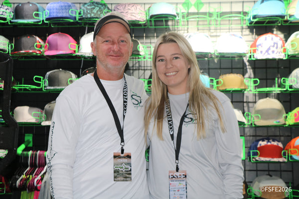 A smiling apparel vendor duo stands in front of a colorful wall of hats at the Florida Fishing Expo.