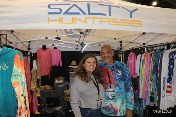 The Salty Huntress vendor duo smile together beneath racks of their vibrant fishing apparel at the Florida Fishing Expo.