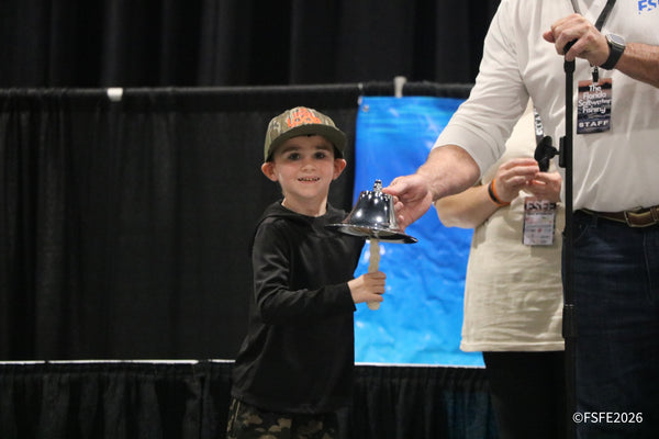 A beaming young attendee rings the ceremonial bell as Florida Fishing Expo staff look on.