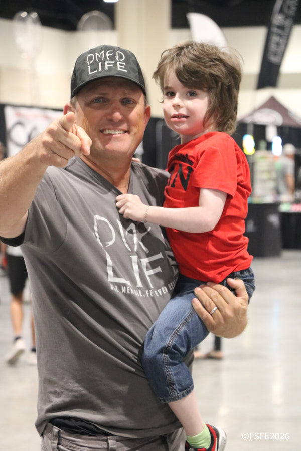 A DMFD Life dad points to the camera while proudly holding his smiling son at the Florida Fishing Expo.