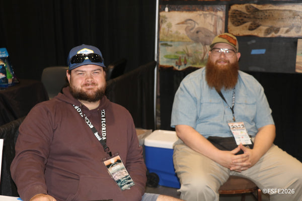 Two vendors relax and smile beside a wildlife-art booth's heron woodburning at the Florida Fishing Expo.