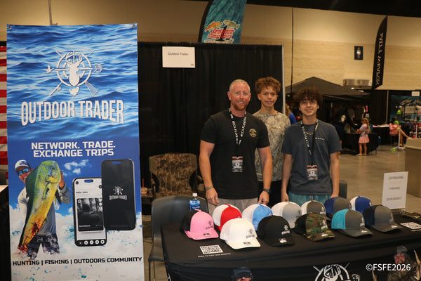 Outdoor Trader's team smiles behind a table of branded caps, promoting their hunting and fishing trip-exchange app at the Florida Fishing Expo.