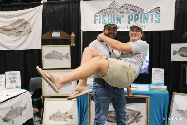 Two laughing vendors goof around at the Fishin' Prints booth, one lifted off his feet, at the Florida Fishing Expo.