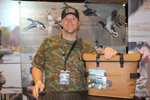 A Dry Pocket vendor smiles beside his waterproof cooler bag, backed by a duck-hunting scene banner at the Florida Fishing Expo.