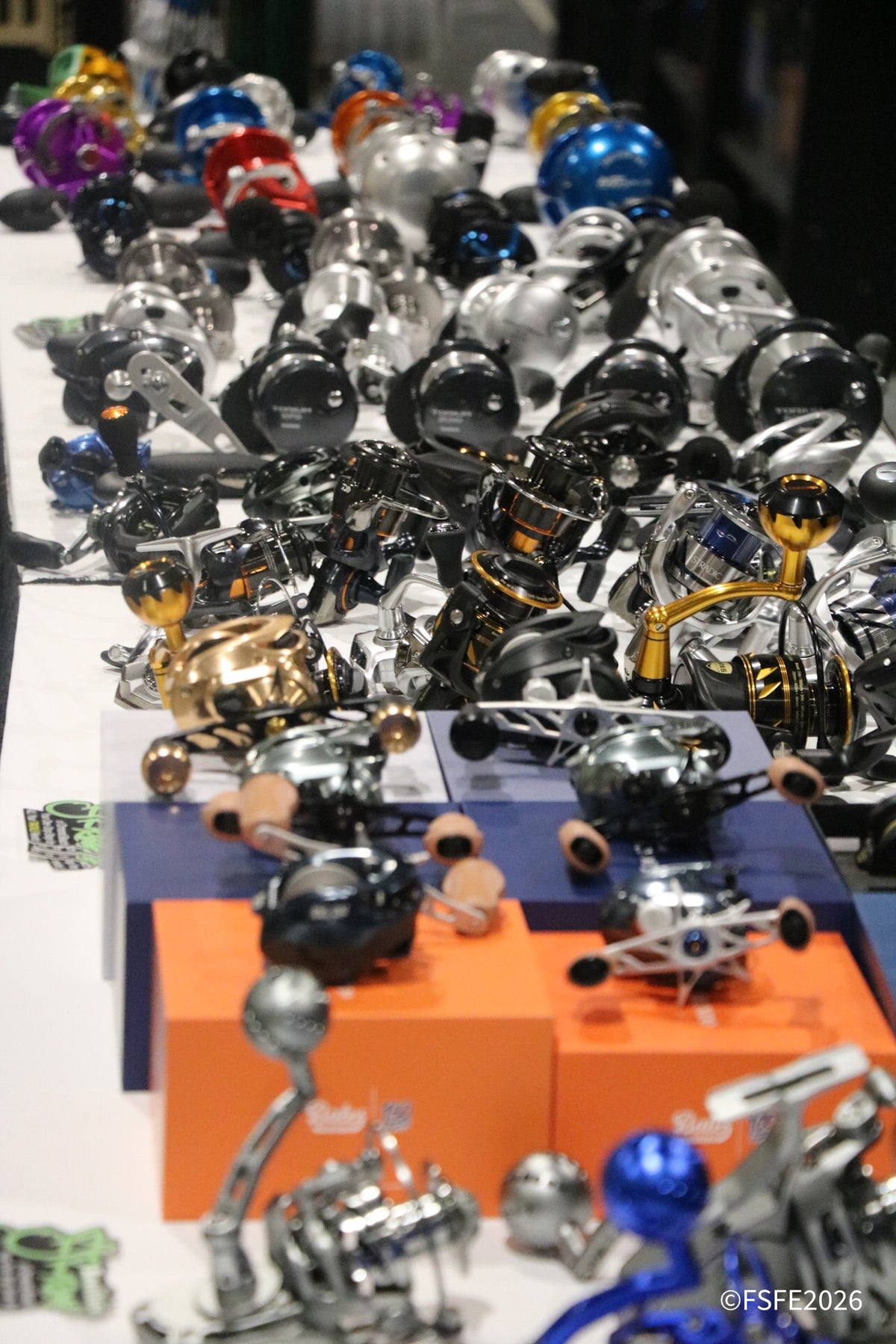 A dense display of fishing reels and related hardware arranged on display stands, including metallic silver, black, blue, gold, and purple models, photographed in a showroom or exhibition setting.